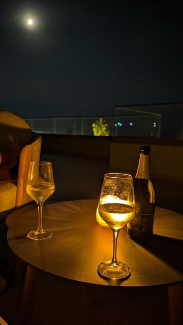 Eine nächtliche Aufnahme auf einer Terrasse zeigt einen kleinen, runden Tisch im Vordergrund, auf dem zwei Weingläser und eine geöffnete Flasche Wein steht. Das rechte Glas wird von einer kleinen, warmen Lichtquelle – vermutlich einer Kerze dahinter – golden und hell erleuchtet, sodass kleine Wassertropfen auf dem Glas funkeln. Im Hintergrund erstreckt sich eine dunkle Terrasse mit einer modernen Glasbrüstung, hinter der in der Ferne eine beleuchtete Palme und vereinzelte Lichter zu sehen sind.…