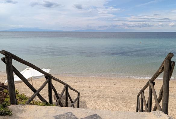 Blick von einer Steintreppe über ein Holzgeländer auf einen menschenleeren Sandstrand und das ruhige, türkisfarbene Meer unter leicht bewölktem Himmel.