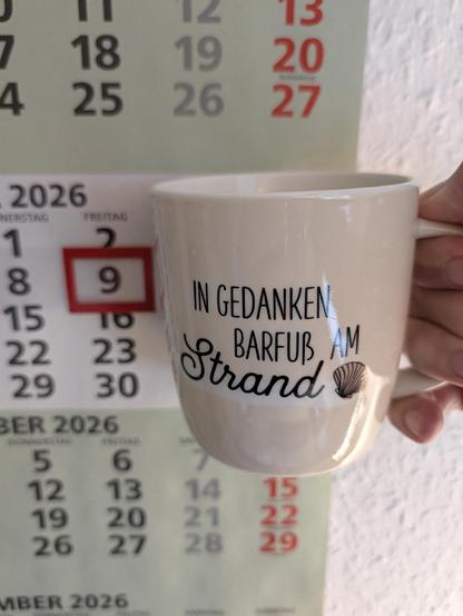 Meine rechte Hand hält einen Porzellan-Kaffeebecher. Seine Farbe ist beige. In schwarzer Schrift steht auf ihm: In Gedanken barfuß am Strand, gefolgt von einer Zeichnung einer bräunlichen Meermuschel. Im Hintergrund hängt ein 3-Monats-Wandkalender an der Wand mit dem rot umrandeten heutigen Tag. 