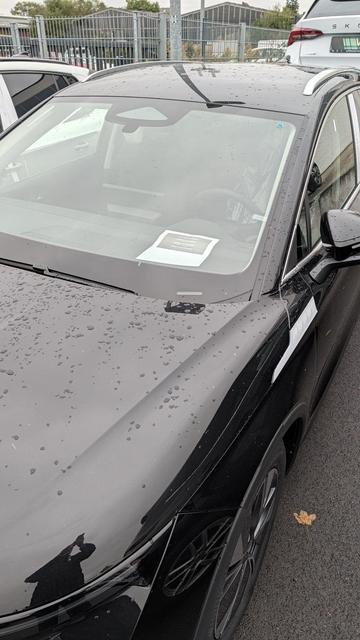 Eine Nahaufnahme von der Frontpartie eines schwarzen, nassgeregneten Autos, das auf einem Parkplatz steht. Auf der glänzenden Motorhaube und der Windschutzscheibe sind zahlreiche Wassertropfen zu sehen. Hinter der Scheibe, auf der Fahrerseite des Armaturenbretts, liegt ein weißes Dokument oder Hinweisschild.