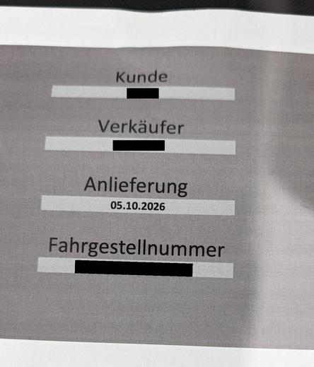 Nahaufnahme des gedruckten Dokuments mit verschiedenen Informationsfeldern auf grauem Hintergrund. Zu sehen sind die zentrierten Beschriftungen „Kunde“, „Verkäufer“, „Anlieferung“ mit dem Datum „05.10.2026“ sowie „Fahrgestellnummer“. Die jeweiligen Eingabefelder darunter sind mit dicken, schwarzen Balken markiert worden, um persönliche Daten unkenntlich zu machen.