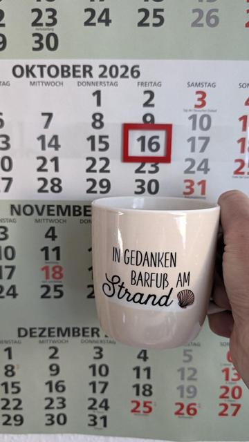 Meine rechte Hand hält einen Porzellan-Kaffeebecher. Seine Farbe ist beige. In schwarzer Schrift steht auf ihm: In Gedanken barfuß am Strand, gefolgt von einer Zeichnung einer bräunlichen Meermuschel. Im Hintergrund hängt ein 3-Monats-Wandkalender an der Wand mit dem rot umrandeten heutigen Tag. 