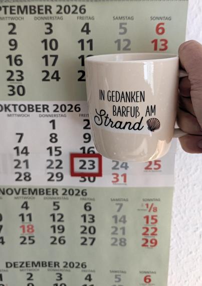 Meine rechte Hand hält einen Porzellan-Kaffeebecher. Seine Farbe ist beige. In schwarzer Schrift steht auf ihm: In Gedanken barfuß am Strand, gefolgt von einer Zeichnung einer bräunlichen Meermuschel. Im Hintergrund hängt ein 3-Monats-Wandkalender an der Wand mit dem rot umrandeten heutigen Tag. 